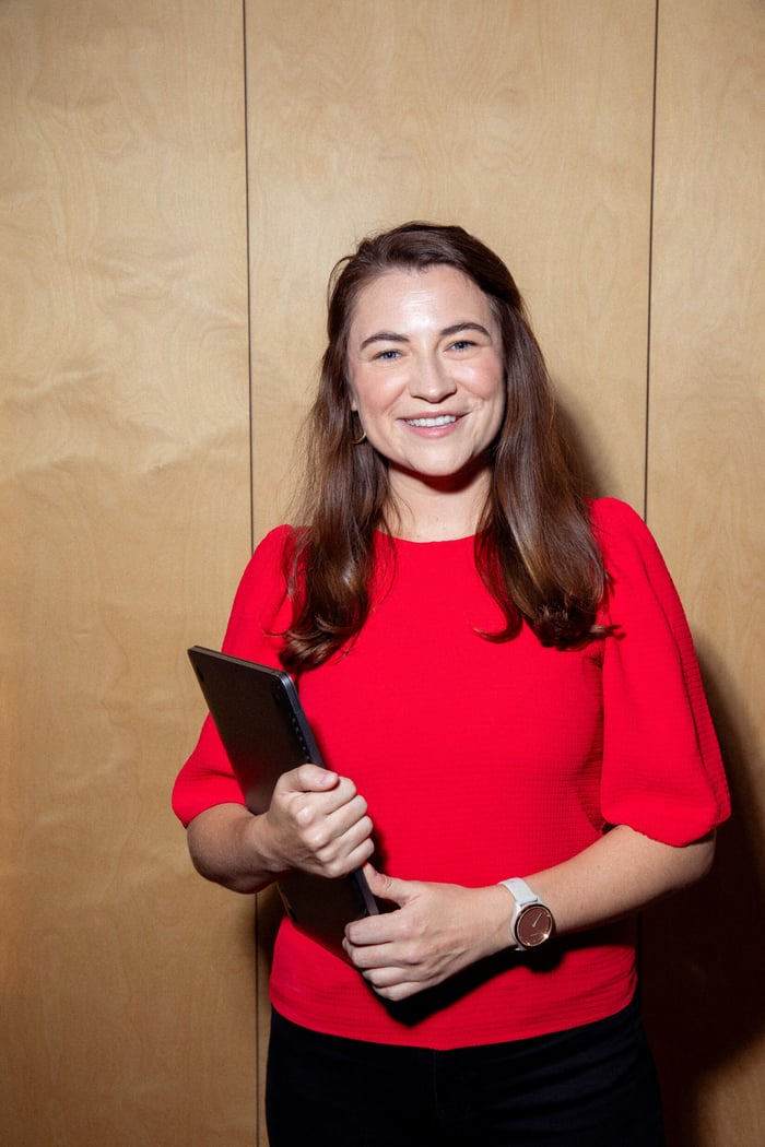 A woman with long brown hair smiles broadly at the camera. She's wearing a bright red top and holding a dark folder or tablet. She's standing in front of a light wooden wall or panel.